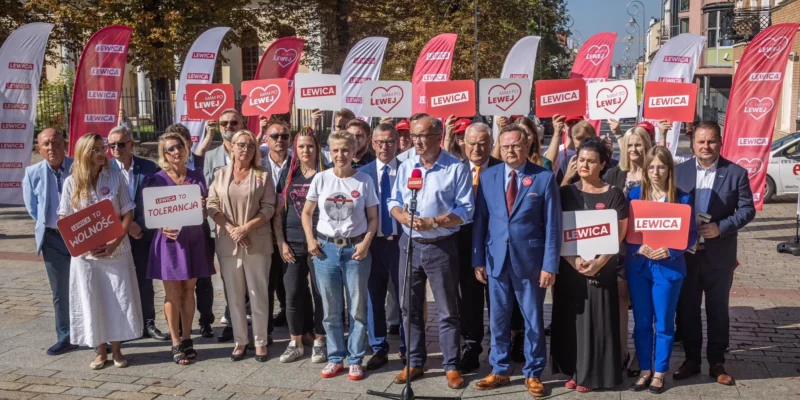 Grupa osób na placu trzyma plakaty i flagi z napisem LEWICA, widoczne hasła „LEWICA TO WOLNOŚĆ” i „LEWICA TO TOLERANCJA” oraz stoi przed mikrofonem.