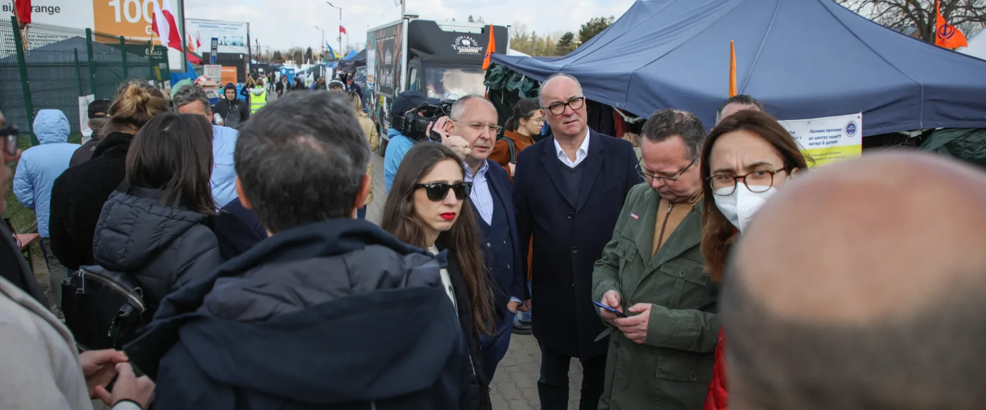 Grupa ludzi w zatłoczonym plenerowym targu z namiotami i straganami, na tle widoczne flagi i banery.