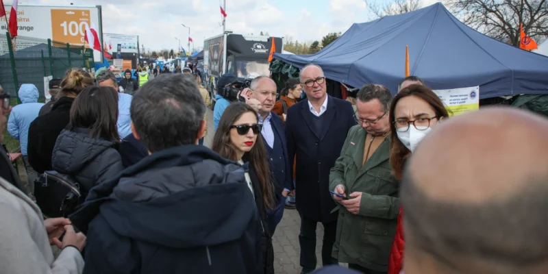 Grupa ludzi w zatłoczonym plenerowym targu z namiotami i straganami, na tle widoczne flagi i banery.