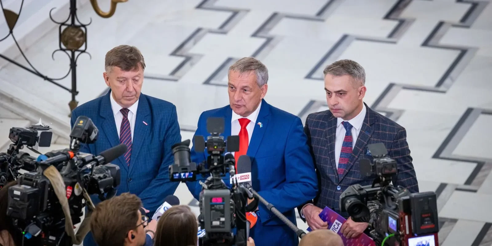 Trzech mężczyzn w garniturach udziela konferencji prasowej otoczonych kamerami, na tle marmurowych schodów i ozdobnego żyrandola, w kontekście apelu o weto.