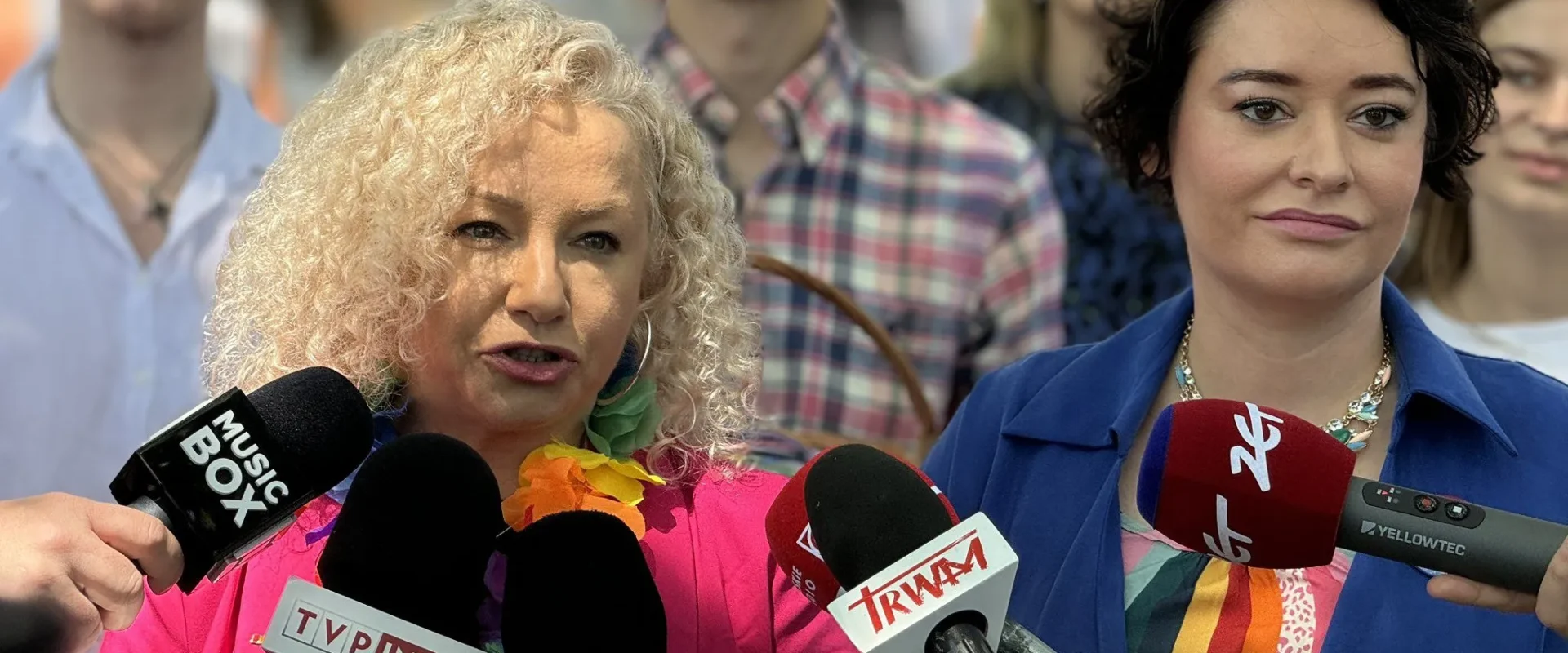 Kobieta z blondymi kręconymi włosami w różowej marynarce przemawia do mikrofonów mediów podczas Parady Równości 2024.