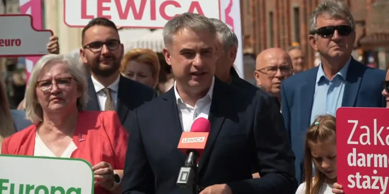 Polityk Lewicy przemawia do mikrofonu podczas plenerowego zgromadzenia, wokół niego ludzie trzymają plakaty i banery z napisem LEWICA.