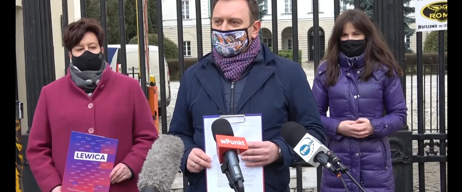 Trzy osoby w maseczkach stojące przed bramą podczas konferencji prasowej Lewicy, trzymające materiały z napisem Lewica i stojące przy mikrofonach.