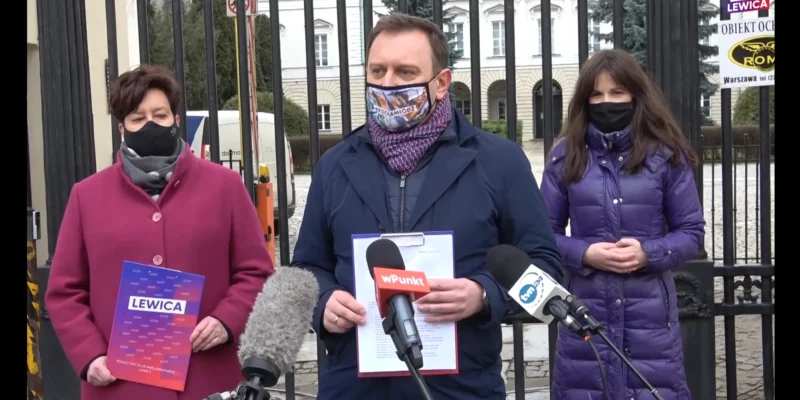 Trzy osoby w maseczkach stojące przed bramą podczas konferencji prasowej Lewicy, trzymające materiały z napisem Lewica i stojące przy mikrofonach.