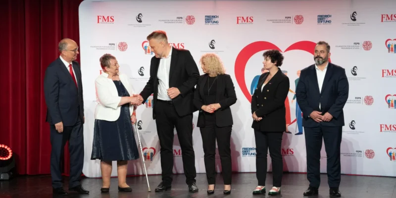 Szóstka osób stoi na scenie podczas wydarzenia; starsza kobieta w białej marynarce ściska dłoń wysokiemu mężczyźnie w czarnym garniturze, a pozostali obserwują, z tyłu biało-czerwony baner z logotypami i dużym czerwonym sercem.