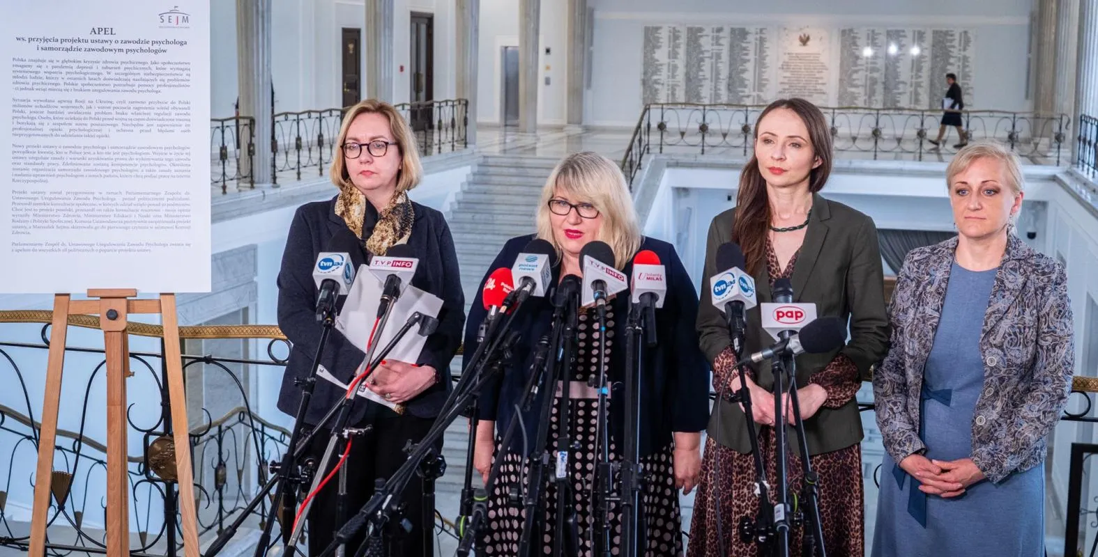 Na zdjęciu przedstawione są cztery kobiety stojące przy mównicy z mikrofonami, podczas konferencji prasowej dotyczącej projektu ustawy o zawodzie psychologa w przestrzeni Sejmu.