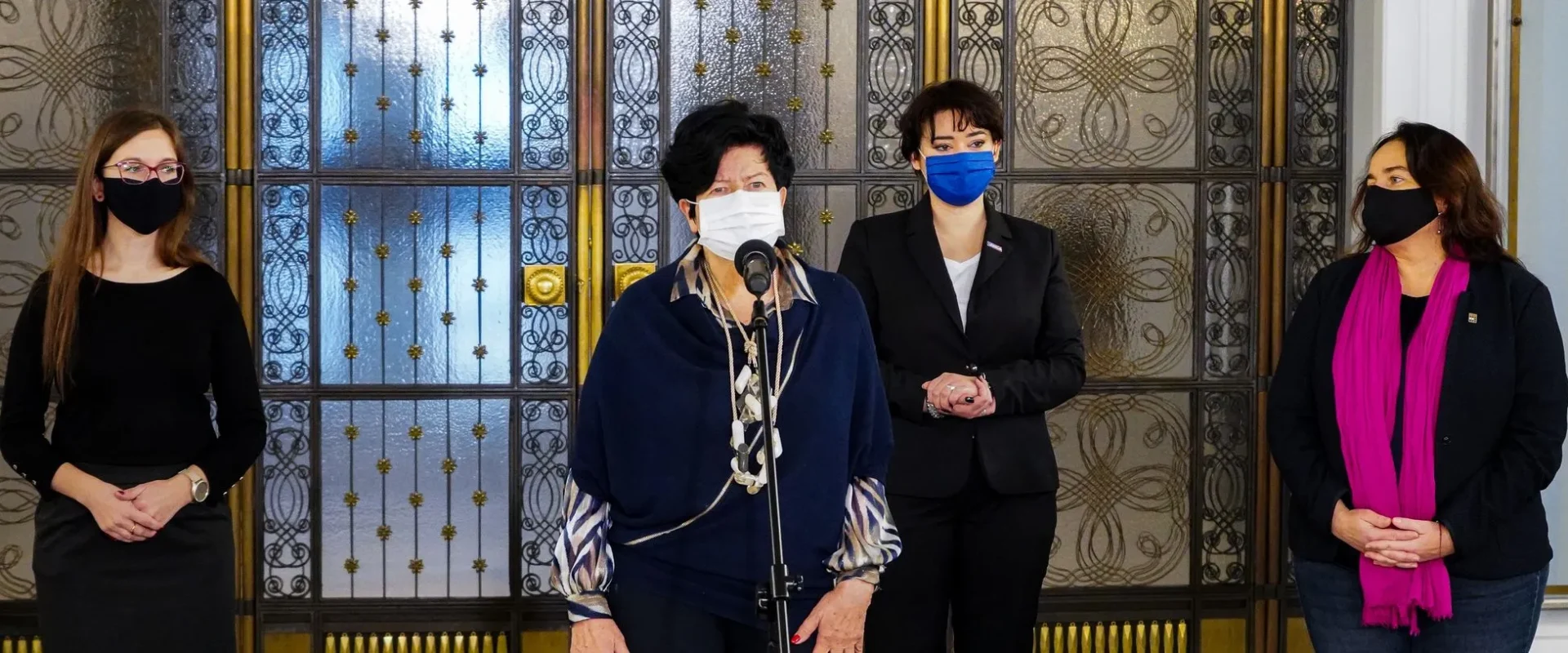 Cztery kobiety w maseczkach stoją przed ozdobnymi metalowymi drzwiami, a kobieta w centrum przemawia do mikrofonu.