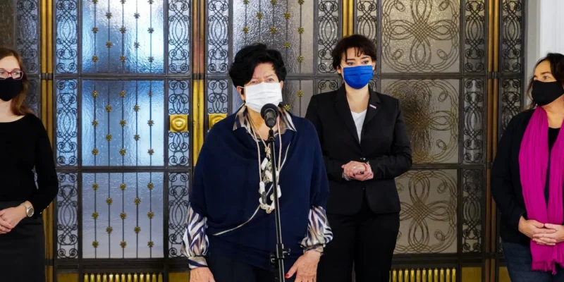 Cztery kobiety w maseczkach stoją przed ozdobnymi metalowymi drzwiami, a kobieta w centrum przemawia do mikrofonu.