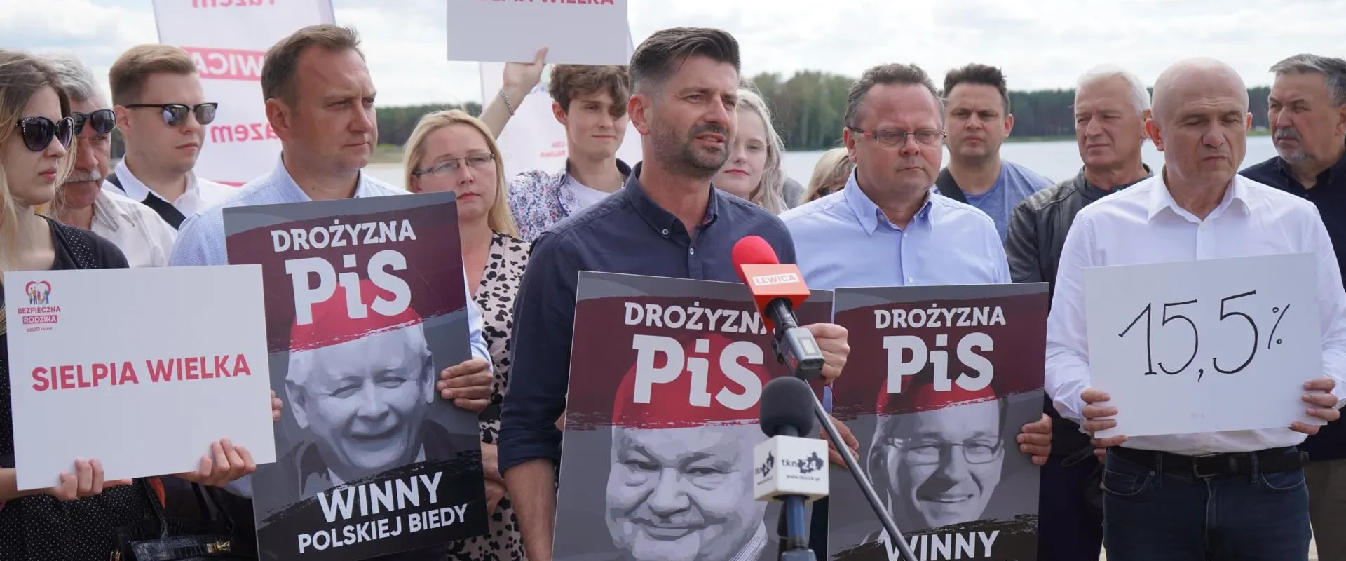 Grupa ludzi na konferencji prasowej nad jeziorem trzyma plakaty z hasłem „DROŻYŻNA PiS” i napisem „WINNY POLSKIEJ BIEDY”, podczas gdy mężczyzna przemawia do mikrofonów.