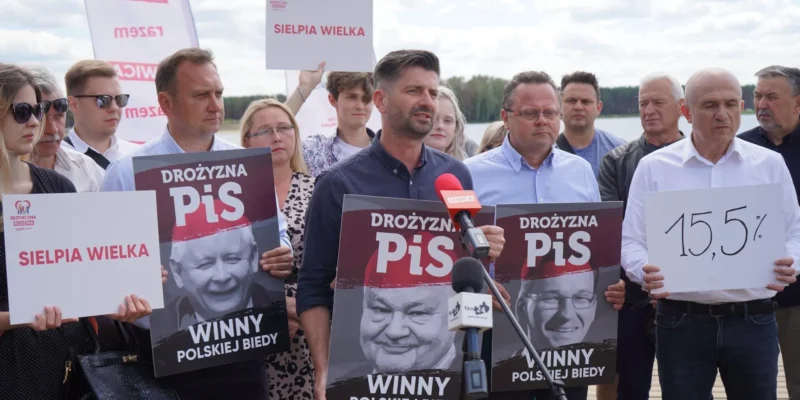 Grupa ludzi na konferencji prasowej nad jeziorem trzyma plakaty z hasłem „DROŻYŻNA PiS” i napisem „WINNY POLSKIEJ BIEDY”, podczas gdy mężczyzna przemawia do mikrofonów.