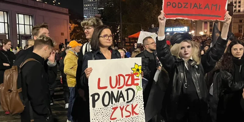 Grupa demonstrantów na wieczornym proteście, jedna osoba trzyma duży plakat „LUDZIE PONAD ZYSK”, druga unosi czerwony baner nad głową; w tle miejskie budynki (Kontekst: Sowińska: Oświadczenie w sprawie Kołodziejczaka).