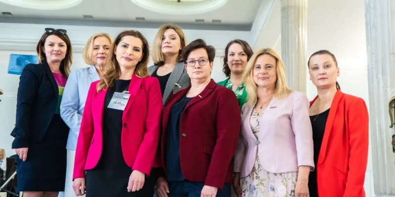 Grupa kobiet w kolorowych marynarkach stoi razem w jasnej sali z kolumnami.