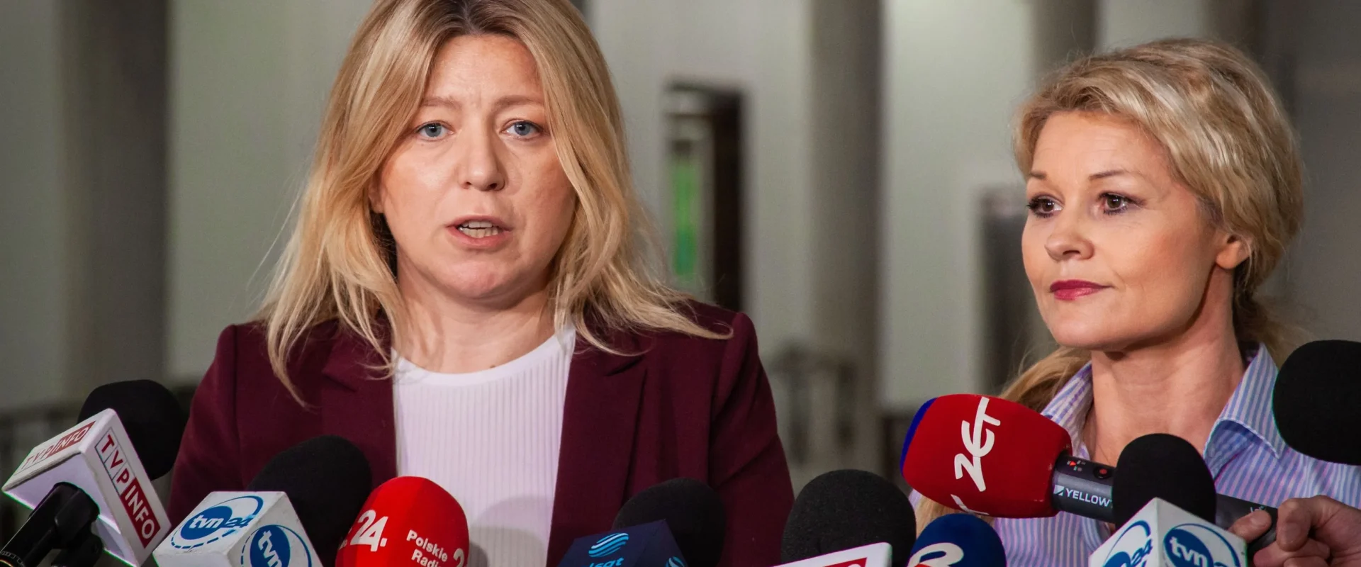Na zdjęciu przedstawione są dwie kobiety przemawiające do mediów, otoczone mikrofonami, w jasnym, biurowym otoczeniu.