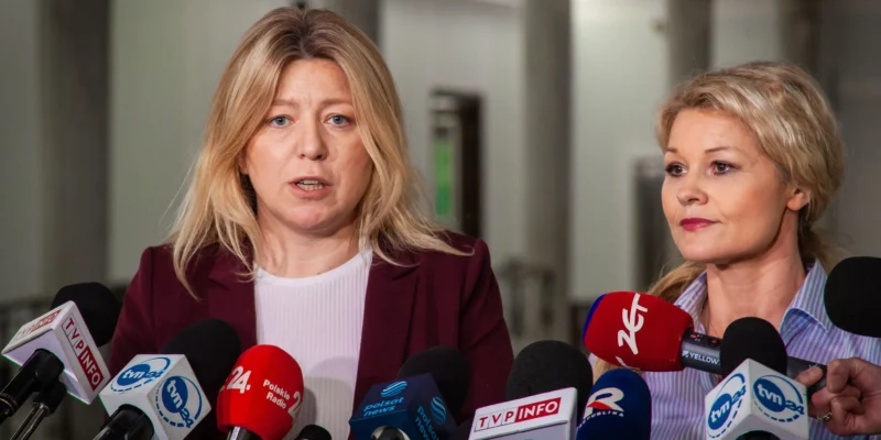 Na zdjęciu przedstawione są dwie kobiety przemawiające do mediów, otoczone mikrofonami, w jasnym, biurowym otoczeniu.