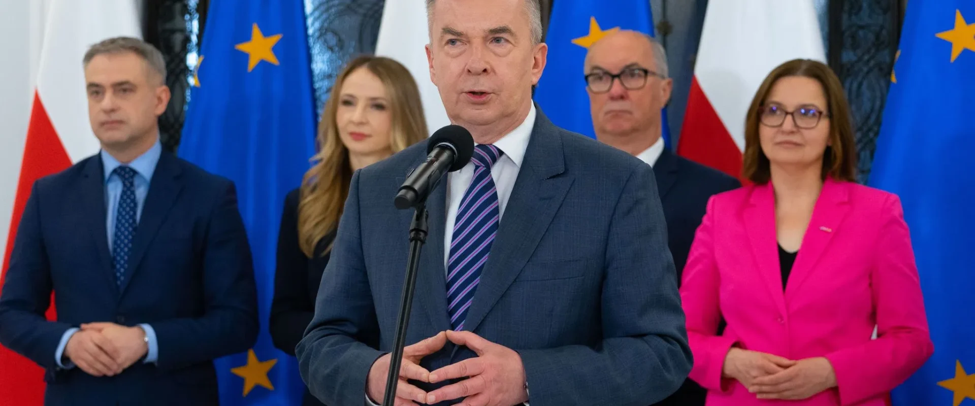 Przedstawiciel Lewicy w rządzie przemawia do mikrofonu podczas konferencji prasowej, na tle stoją flagi Polski i Unii Europejskiej oraz grupa współpracowników.