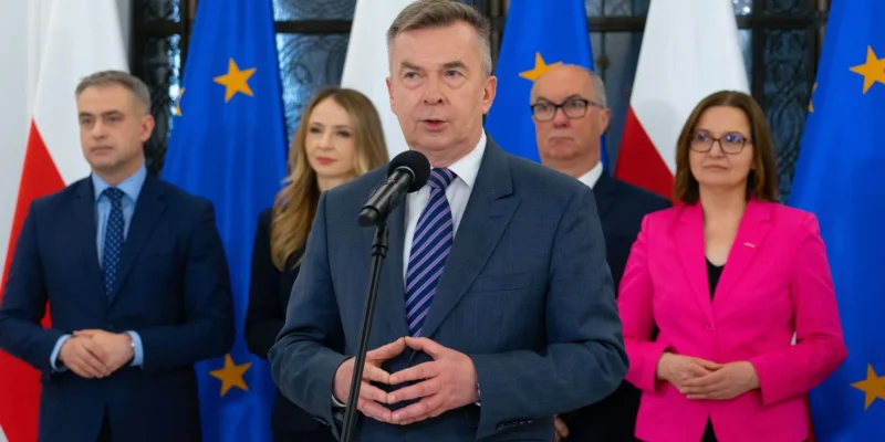 Przedstawiciel Lewicy w rządzie przemawia do mikrofonu podczas konferencji prasowej, na tle stoją flagi Polski i Unii Europejskiej oraz grupa współpracowników.