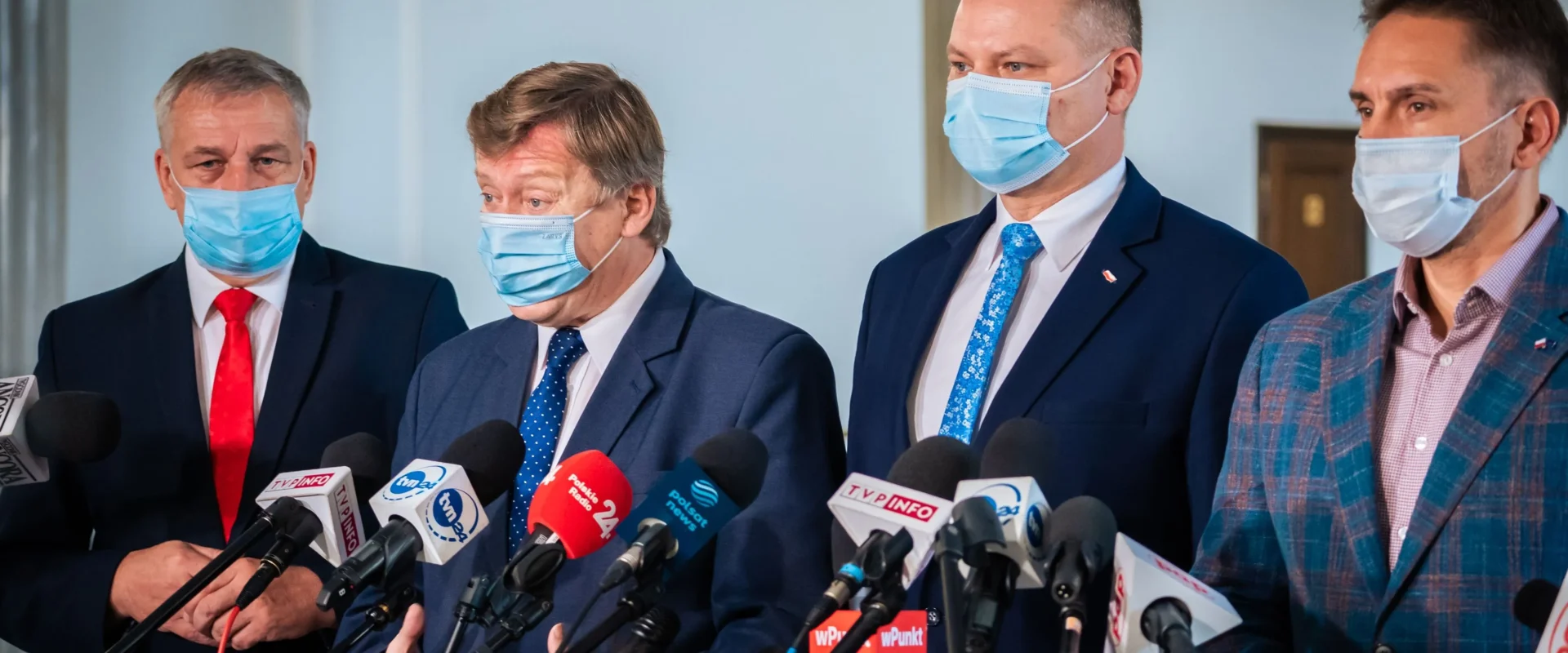 Czterech mężczyzn w garniturach i maseczkach stoją przed licznymi mikrofonami podczas konferencji prasowej.