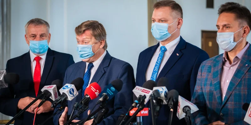 Czterech mężczyzn w garniturach i maseczkach stoją przed licznymi mikrofonami podczas konferencji prasowej.