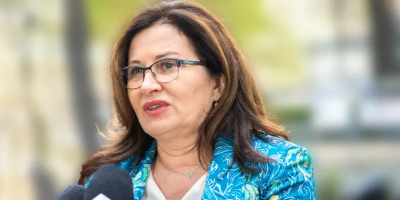 Kobieta w okularach, ubrana w niebieski wzorzysty żakiet, przemawia do mikrofonu podczas wywiadu.