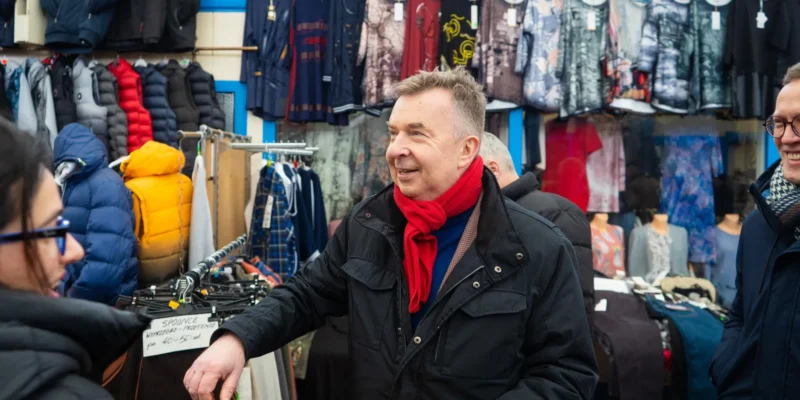 Uśmiechnięty mężczyzna w czarnym płaszczu i czerwonym szaliku rozmawia z innymi klientami na stoisku z odzieżą na targu w Zachodniopomorskiem.