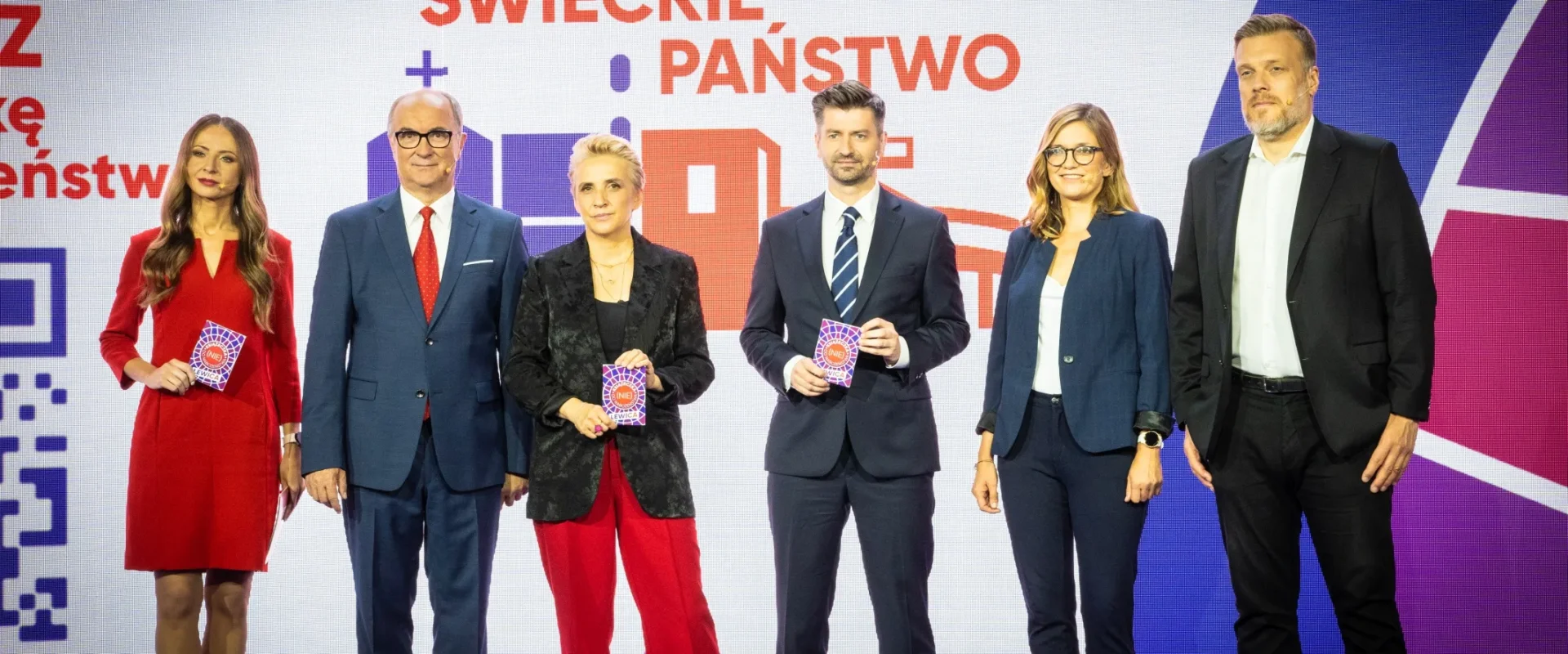 Sześć osób w eleganckich strojach stoi na scenie, trzymając kolorowe karty, a tło stanowi duży ekran z napisem „Świeckie państwo – Teraz!”.
