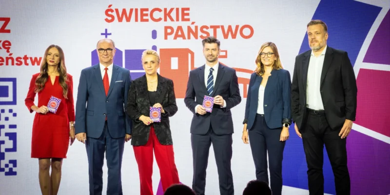 Sześć osób w eleganckich strojach stoi na scenie, trzymając kolorowe karty, a tło stanowi duży ekran z napisem „Świeckie państwo – Teraz!”.