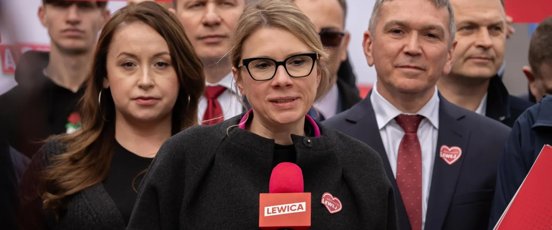 Kobieta w czarnym płaszu i czarnych okularach przemawia do mikrofonu z napisem Lewica, stojąc przed grupą zwolenników z czerwonymi plakata w tle.