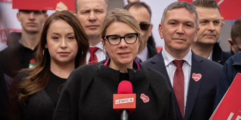 Kobieta w czarnym płaszu i czarnych okularach przemawia do mikrofonu z napisem Lewica, stojąc przed grupą zwolenników z czerwonymi plakata w tle.