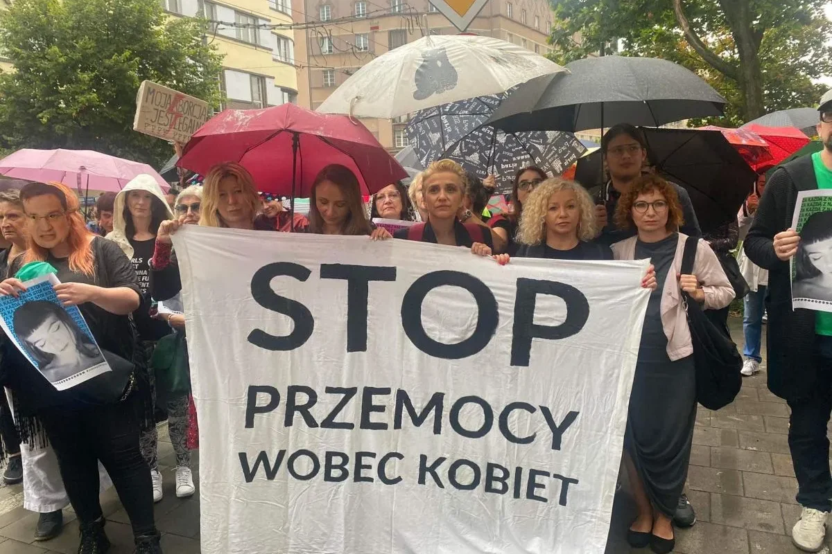 Grupa ludzi na demonstracji trzyma duży biały banner z napisem STOP PRZEMOCY WOBEC KOBIET i stoi pod parasolkami na ulicy.