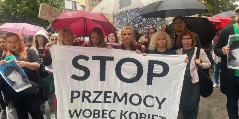 Grupa ludzi na demonstracji trzyma duży biały banner z napisem STOP PRZEMOCY WOBEC KOBIET i stoi pod parasolkami na ulicy.