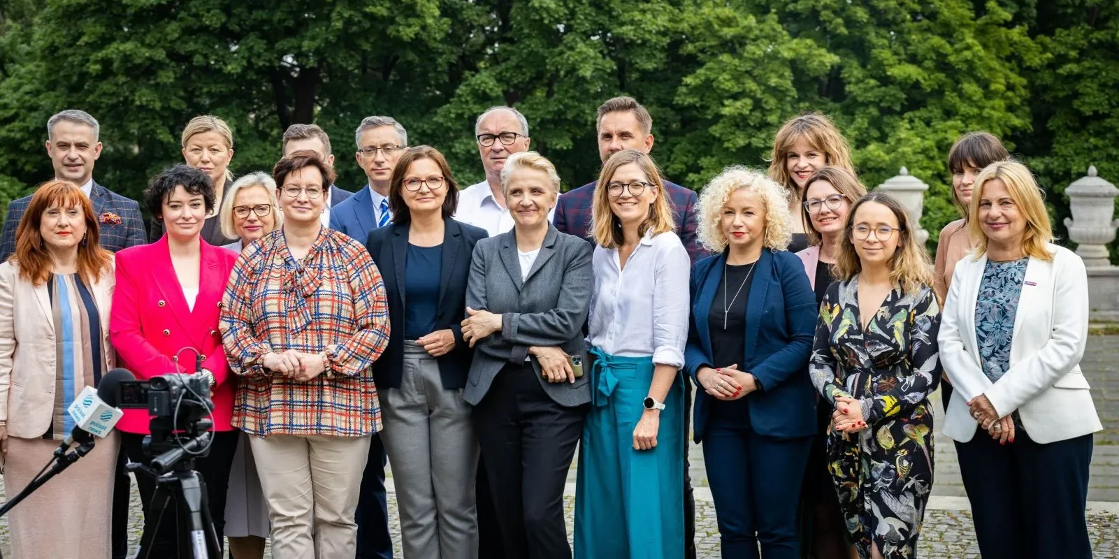 Grupa osób w eleganckich ubraniach stoi razem na zewnątrz, na tle zielonego parku, podczas konwencji „Mamy plan dla mam”.