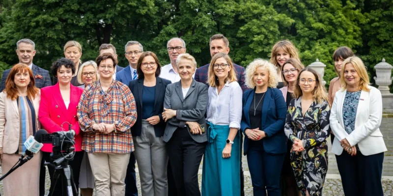 Grupa osób w eleganckich ubraniach stoi razem na zewnątrz, na tle zielonego parku, podczas konwencji „Mamy plan dla mam”.