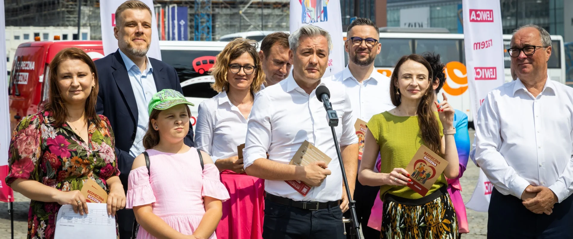 Grupa osób na konferencji prasowej na zewnątrz, mężczyzna w białej koszuli przemawia do mikrofonu i trzyma broszury, wokół niego stoją inni uczestnicy i dziecko; w tle widoczne banery i zabudowania.