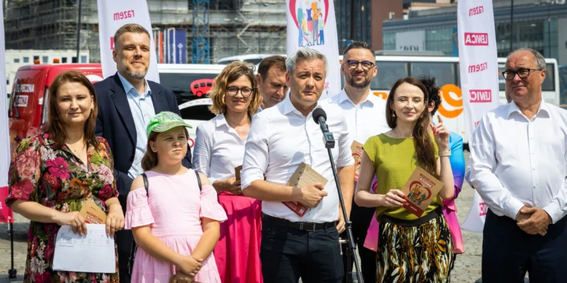 Grupa osób na konferencji prasowej na zewnątrz, mężczyzna w białej koszuli przemawia do mikrofonu i trzyma broszury, wokół niego stoją inni uczestnicy i dziecko; w tle widoczne banery i zabudowania.