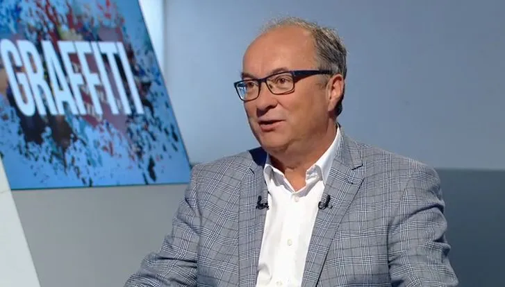 Mężczyzna w okularach, ubrany w jasnoszarą kratowaną marynarkę i białą koszulę, siedzi w studiu telewizyjnym podczas wywiadu, a w tle znajduje się ekran z napisem GRAFFITI.