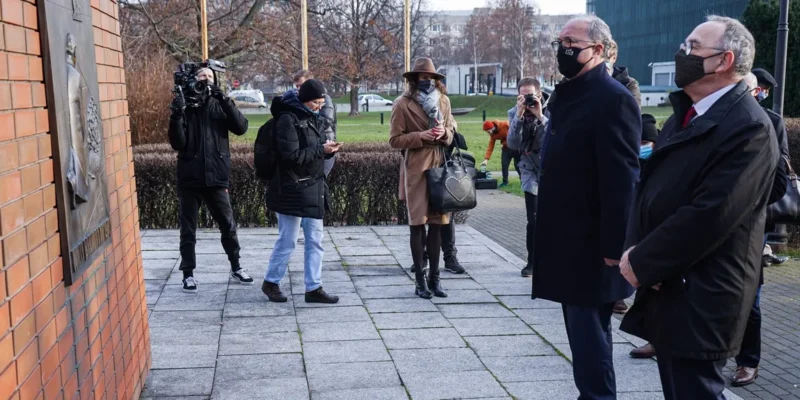 Grupa ludzi z aparatami i dziennikarzami stoi na zewnątrz przy tablicy upamiętniającej gest Willy’ego Brandta.