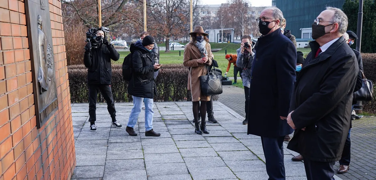 Grupa ludzi z aparatami i dziennikarzami stoi na zewnątrz przy tablicy upamiętniającej gest Willy’ego Brandta.