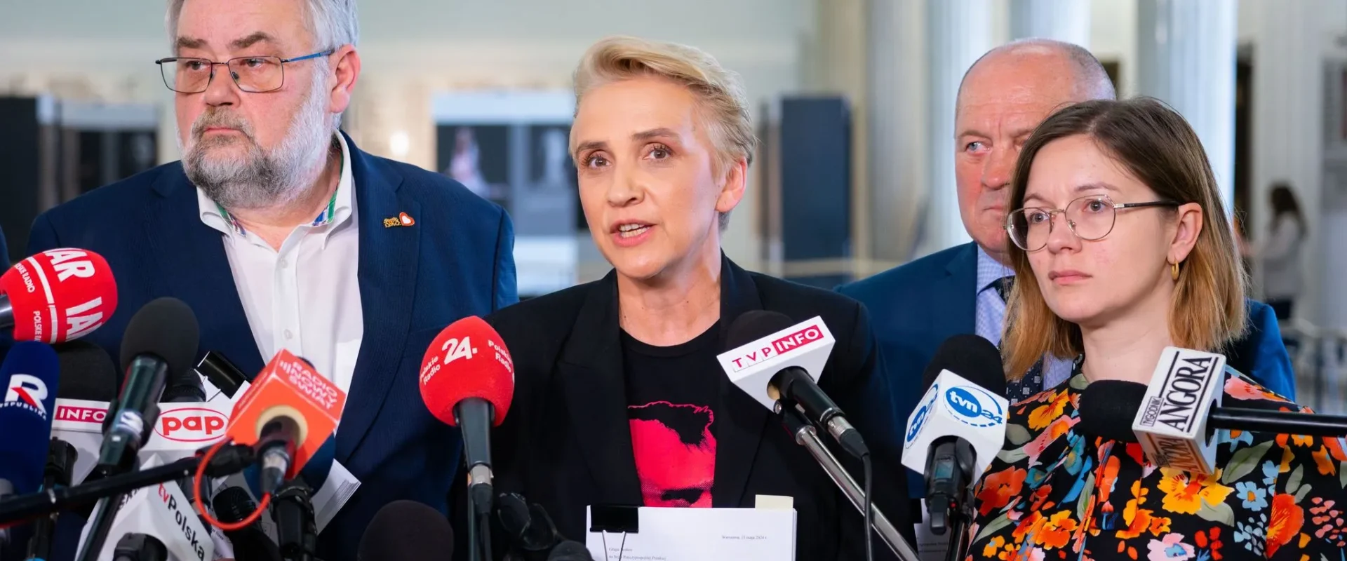 Konferencja prasowa: kobieta w czarnym żakiecie przemawia do licznych mikrofonów, stojąc między dwoma mężczyznami i trzymając dokument.