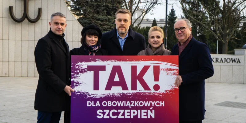 Pięć osób stoi na placu i trzyma duży gradientowy plakat z napisem TAK! DLA OBOWIĄZKOWYCH SZCZEPIEŃ (LEWICA), na tle widoczny pomnik i przestrzeń miejska.