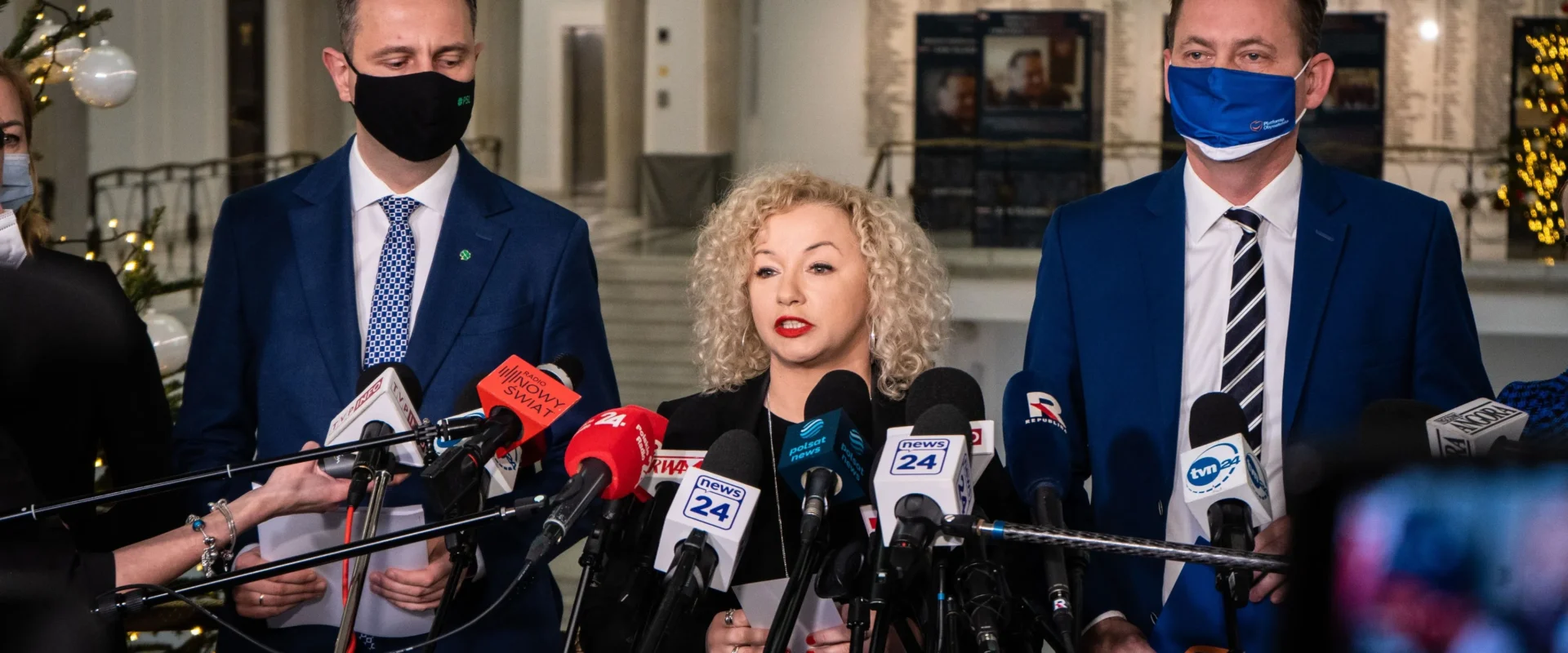 Trójka polityków w garniturach udziela konferencji prasowej, otoczona mikrofonami, wszyscy w maseczkach, w reprezentacyjnym wnętrzu Sejmu.