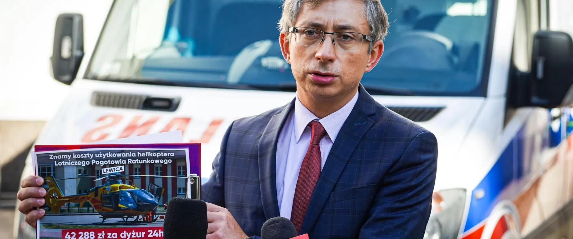 Mężczyzna w garniturze z czerwonym krawatem stoi przy mikrofonach, trzyma plakat z informacją o kosztach lotu helikoptera Lotniczego Pogotowia Ratunkowego, na tle pojazdu ratunkowego.