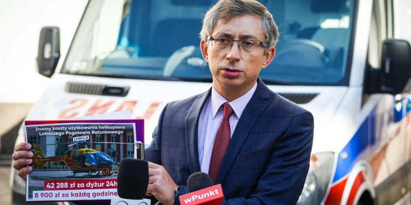 Mężczyzna w garniturze z czerwonym krawatem stoi przy mikrofonach, trzyma plakat z informacją o kosztach lotu helikoptera Lotniczego Pogotowia Ratunkowego, na tle pojazdu ratunkowego.