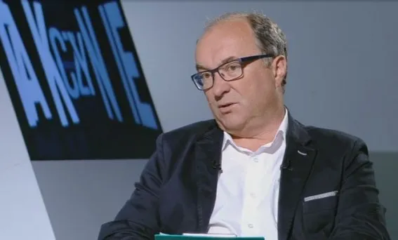 Mężczyzna w okularach i marynarce siedzi w studiu telewizyjnym, trzymając dokumenty podczas rozmowy.