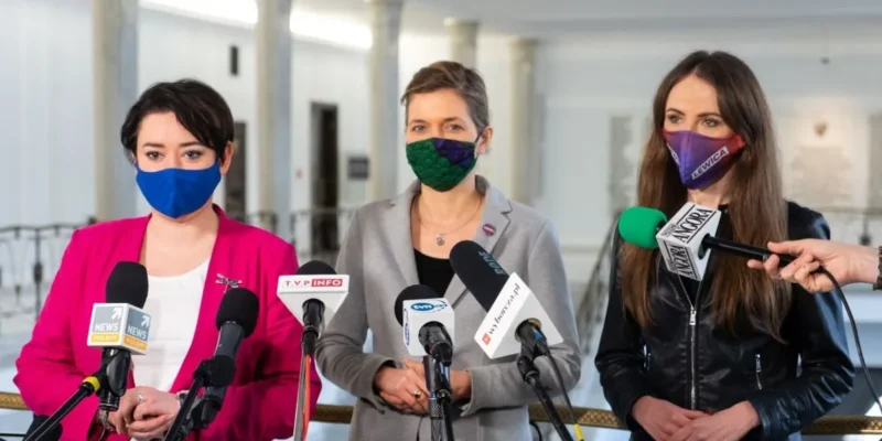 Trzy kobiety stojące przy mikrofonach podczas konferencji prasowej w jasnym holu, każda ma inną maseczkę: niebieską, zieloną i fioletową.