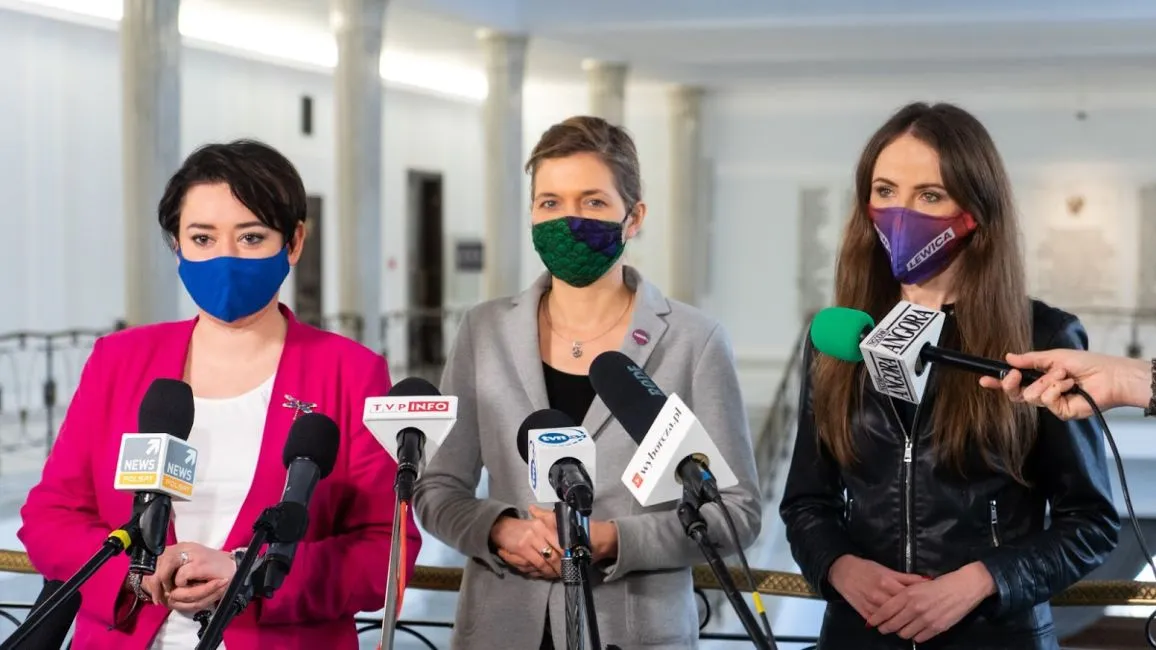 Trzy kobiety stojące przy mikrofonach podczas konferencji prasowej w jasnym holu, każda ma inną maseczkę: niebieską, zieloną i fioletową.