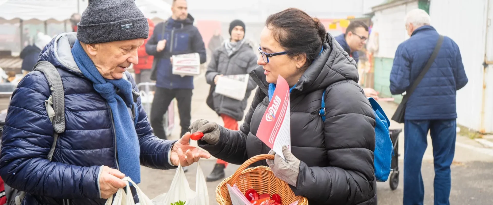 Na zdjęciu widoczna jest interakcja między starszym mężczyzną a kobietą, która trzyma koszyk, podczas zakupów na rynku w mglisty dzień.