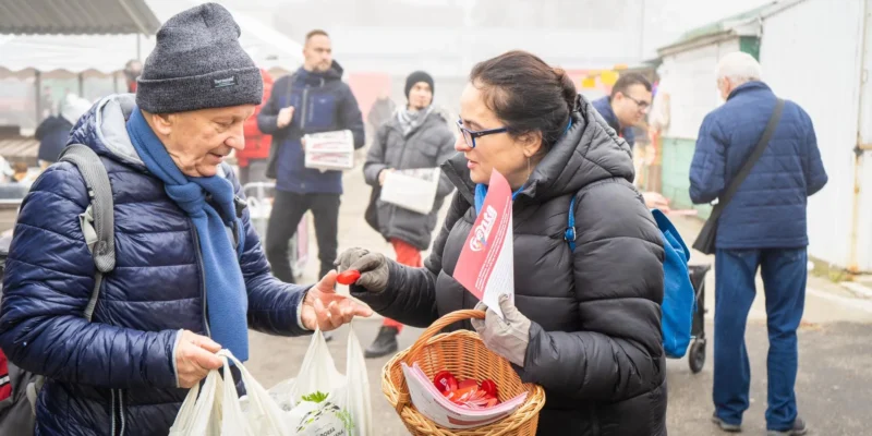 Na zdjęciu widoczna jest interakcja między starszym mężczyzną a kobietą, która trzyma koszyk, podczas zakupów na rynku w mglisty dzień.