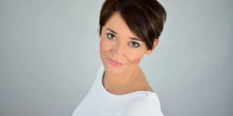 Portret kobiety o krótkich brązowych włosach, delikatnie uśmiechającej się do kamery na jasnym tle, ubranej w białą bluzkę.