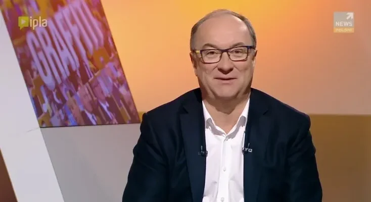 Uśmiechnięty mężczyzna w okularach i ciemnej marynarce siedzi w studiu telewizyjnym.