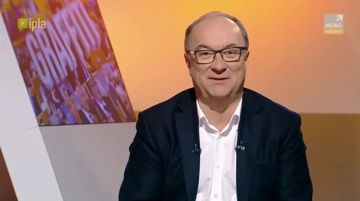 Uśmiechnięty mężczyzna w okularach i ciemnej marynarce siedzi w studiu telewizyjnym.